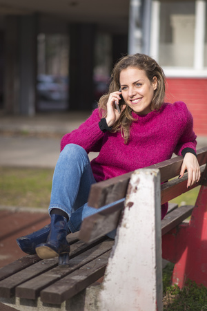 Sweet young woman sitting on the bench in a park, having a wonderful conversation on her phone, feeling free and relaxedの写真素材