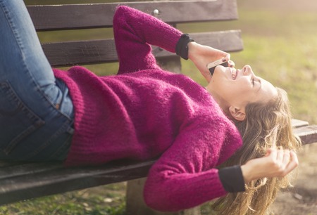 Sweet young woman laying down on the bench in a park, having a wonderful conversation on her phone, feeling free and relaxedの写真素材
