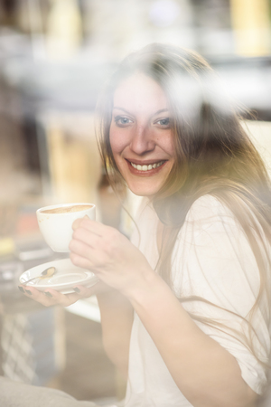 Cheerful young casual lady drinking her coffee in a coffee shop, enjoying some free time for herself, through the window shotの写真素材