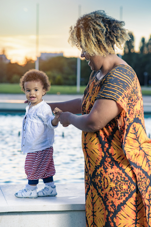 Young african american mother teaching her baby daughter how to walk near a fountain during a wonderful sunsetの写真素材