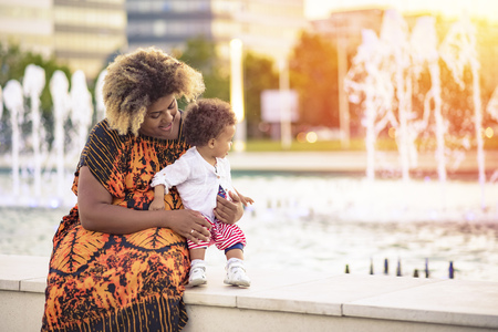 Young african american mother teaching her baby daughter how to walk near a fountain during a wonderful sunsetの写真素材