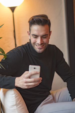 Young attractive man typing a chat message on his cellphone, sitting in a coffee shopの写真素材