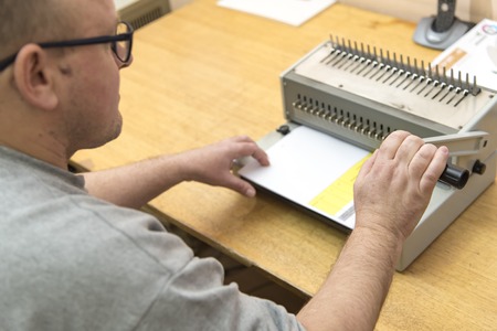 Young man using a manual paper binder to bind documents, stack of papers, together