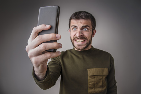 Young man angry and furious with his cell phone, shouting and yelling in shockの写真素材
