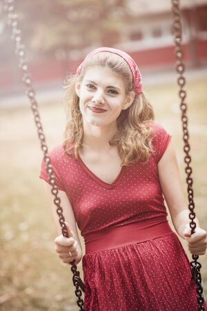 Happy young woman in red retro dress swinging on a swing in a park on a sunny dayの写真素材