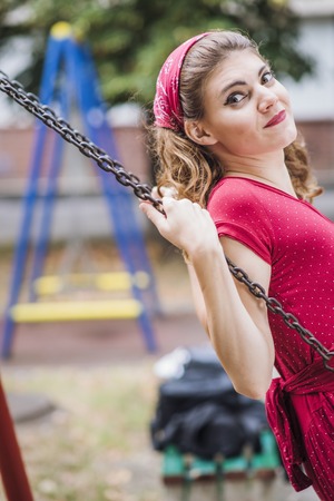 Happy young woman in red retro dress swinging on a swing in a park on a sunny dayの写真素材