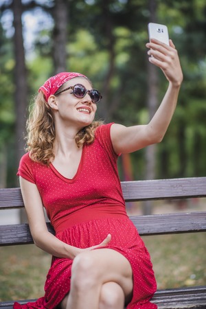 Young woman in a red retro dress taking a selfie, posing in front of her cell phone cameraの写真素材