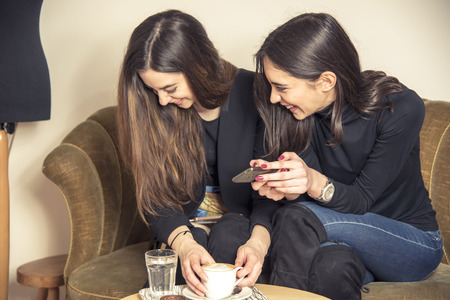 Two beautiful girl friends sitting in a coffee shop, drinking coffee, using cell phones and having a bit of gossipの写真素材
