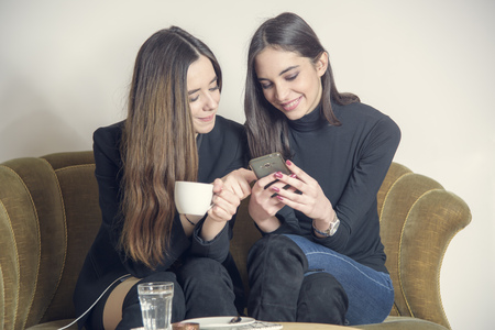Two beautiful girl friends sitting in a coffee shop, drinking coffee, using cell phones and having a bit of gossipの写真素材