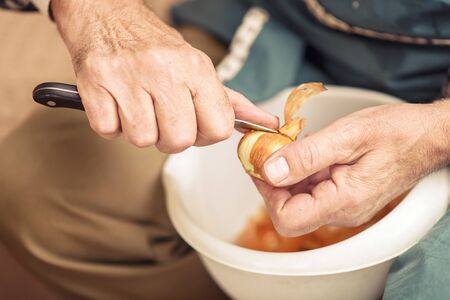 Senior elderly man peeling onion skin with a small sharp knife, preparing ingredients for lunchの写真素材