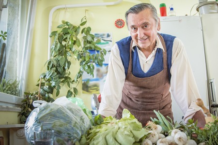Senior old man in apron in his kitchen, standing over vegetables and smilingの写真素材