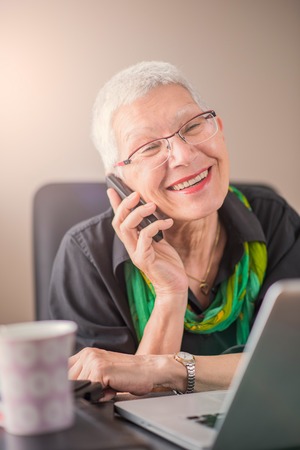 Happy senior business woman talking on her cell phone, having a lovely business chat in her officeの写真素材