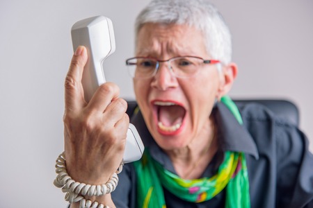 Angry, enraged senior woman yelling at a landline office phone, unhappy with customer service provided by the agent on the other sideの写真素材