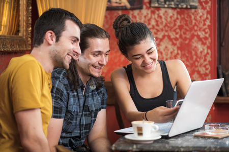 Group of friends, young people, sitting in a coffee shop, having fun using a laptop and a phoneの写真素材