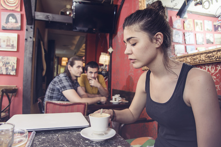 Two young men gossiping and talking behind a girls back in a coffee shopの写真素材