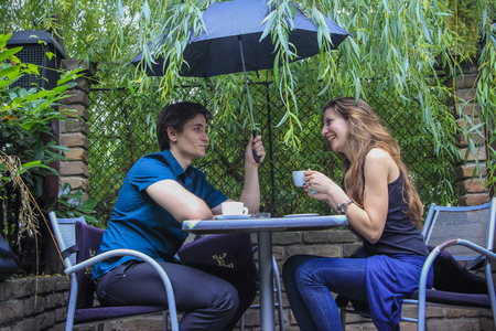 Cute couple enjoying a cup of espresso coffee on a rainy day in an outdoor coffee shop, gentleman holding the umbrellaの写真素材