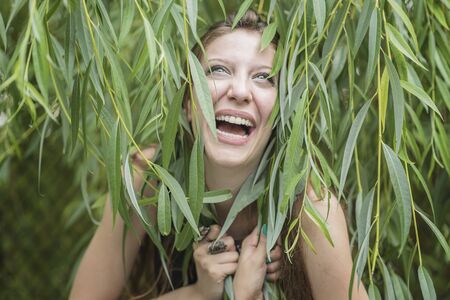 Cheerful young beautiful woman smiling through vibrant green willow branches, enjoying nature and sunny dayの写真素材