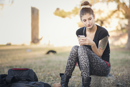 Young alternative fashion girl typing a message on her cell phone, sitting in the parkの写真素材