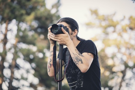 Young photographer with long hair and alternative style taking photographs with his dslr camera, capturing landscape and sunset in a parkの写真素材