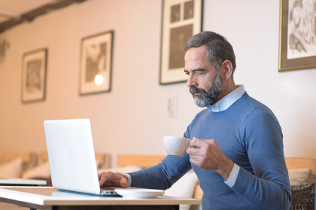 Casual senior man sitting in a coffee shop, having a coffee while browsing internet on his laptopの写真素材
