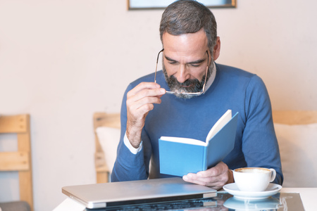 Senior man having troubles reading a book with his glasses, coffee shop surroundingの写真素材