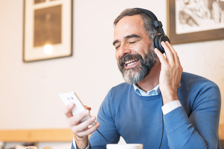 Senior old man listening to his favorite music through big headphones, enjoying the rich sound of the music, hes sitting in a coffee shopの写真素材