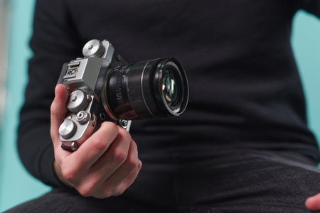 A person is seated, holding a modern camera with a lens while dressed in casual attire. The...