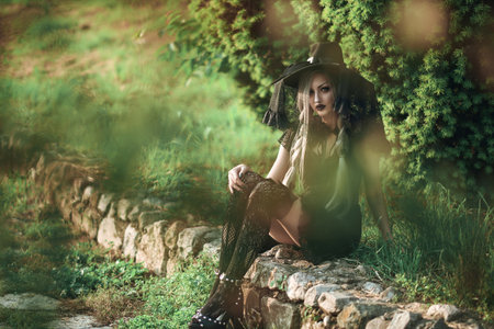 A young gothic woman dressed in a witch costume rests on a stone path, surrounded by lush green...