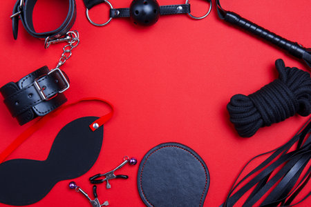 BDSM toys for sex and punishment in the form of a frame. Obedience and authority of man and woman on a red background. Lash, handcuffs, bells, mask, rope and leather spanking.の写真素材