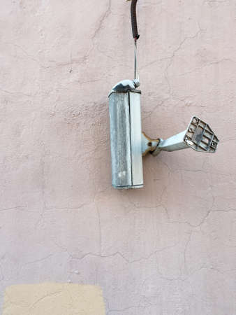 The surveillance camera came off and hangs on the wire. Hooligans broke and filming does not work. Against the background of a gray and brown wall. Old and dirty applianceの写真素材