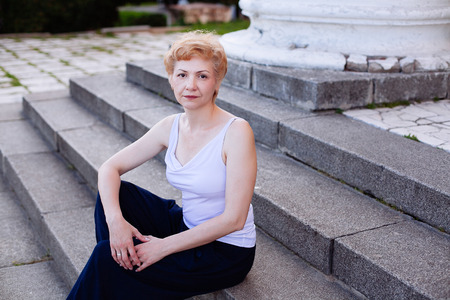 Portrait confident attractive middle aged woman sitting relaxed and happy smiling outdoor on steps in parkの写真素材