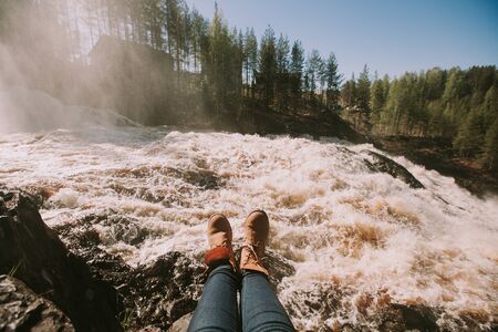 Woman's legs in blue jeans and light  brown boots sitting on the edge near waterfall. Copy spaceの写真素材
