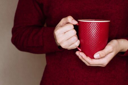Young woman in red sweater is holding red cup of tea or coffee. Trying to warm hands with hot drink