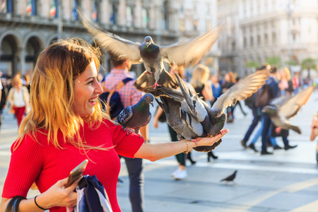 MILAN, ITALY - 17 APRIL 2018 - Unidentified European woman smiles while feeding pigeons with some grain on her hand at the piazza in front of Duomo di Milano.のeditorial素材