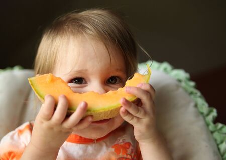 Young baby girl sitting in a high chair eating a wedge of cantaloupe with juice dripping off her chin.の写真素材