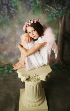 Beautiful young girl wearing pink angel wings and flower halo sitting on a pedestalの写真素材