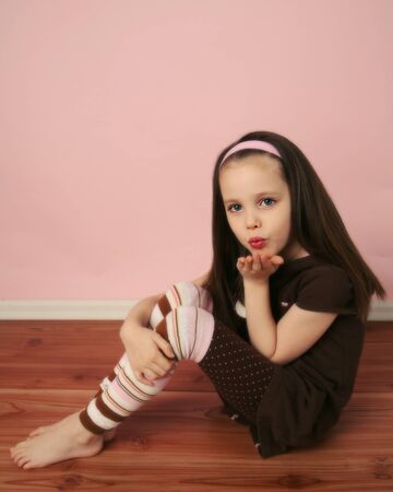 Cute young girl sitting on a wood floor with pink background wall, blowing a kissの写真素材