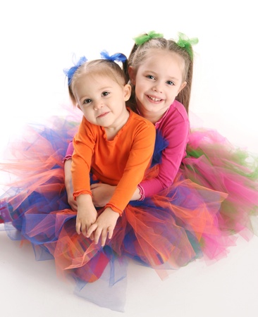 Two adorable sisters dressed in bright colorful tutus sitting down hugging each other, isolated on whiteの写真素材