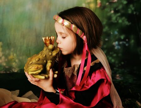 Portrait of a cute young preschool girl dressed as a princess in a pink and gold gown, posing and kissing a frog prince wearing a crownの写真素材