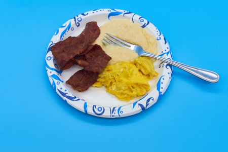 breakfast turkey bacon grits and scrambled eggs on paper plate and light blue backgroundの写真素材