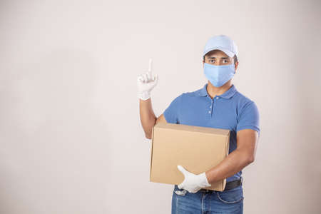 Mexican Latin delivery man carrying a box pointing up wearing a face mask due to covid 19 pandemicの写真素材