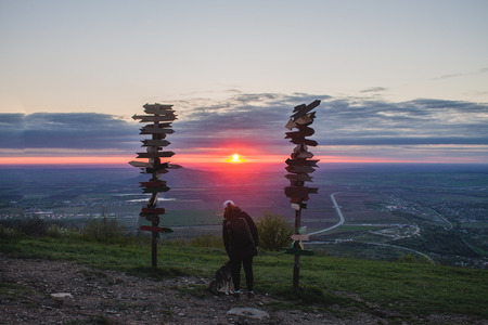 Girl stroking a dog on top of a mountain looking at the sunset and the plain, next to the wooden signs of the cities.の写真素材