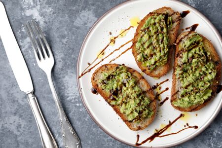 A freshly made minced avocado toast with balsamico dressing, linseed or flaxseed and oil on a white plate, on a gray background and silver fork and knifeの写真素材