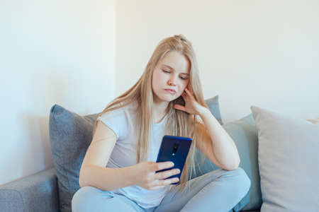 Bored young girl procrastinating and using a smart phone and sitting on a grey couch in the living roomの写真素材