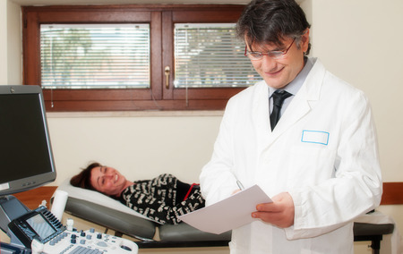 European man smiling doctor visit for medical office patient woman on the medical bed. Writes medical record medical test results.の写真素材