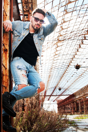 young man on a ladder with denim jacket, black shirt and pants of low waist jeans. metropolitan background. Sunglasses in one hand. a hand through his hair.の写真素材