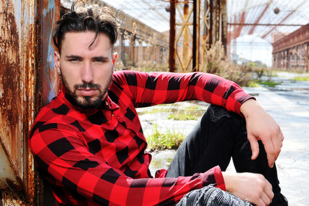 young man sitting on the ground with urban background. He is wearing a red shirt and black pantsの写真素材