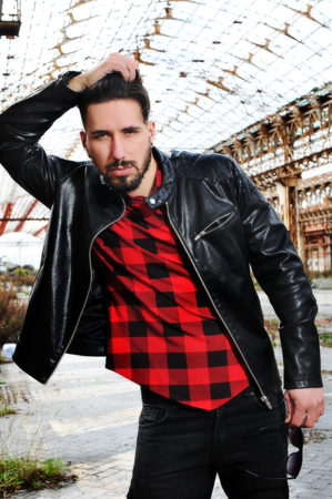 young trendy man with leather jacket, red shirt and black pants with low waist. metropolitan background. Sunglasses in one hand. a hand in the hairの写真素材