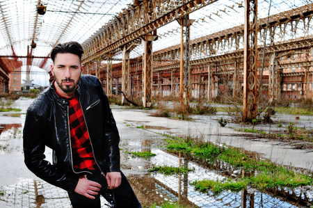 young trendy man with leather jacket, red shirt and black pants with low waist. metropolitan background. Sun glasses in handの写真素材