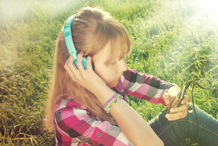 young beautiful girl with headphones on ears listening music in smartphone on the meadow in vintage styleの写真素材